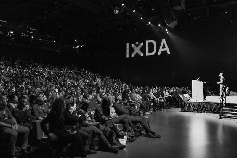 Audience in the auditorium for Interaction 19. There is an IxDA logo on the wall, and the speaker is on the stage to the right of the photo